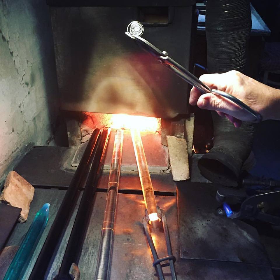 Glasknap holdes i en tang, hvor der bagved er en lille oven hvor tre glas rør bliver opvarmet. 